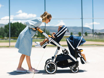 A photo of a Double Stroller by OleOle with a happy Mom and her children. It is a versatile stroller that can be used as a single or double stroller. It is suitable for babies from 6 months to 3 years old. The stroller has a reclining seat.