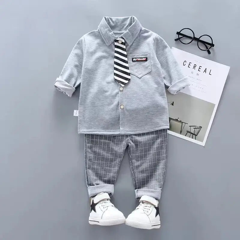 Children's outfit with gray shirt, striped tie, plaid pants, and white sneakers on a gray background.