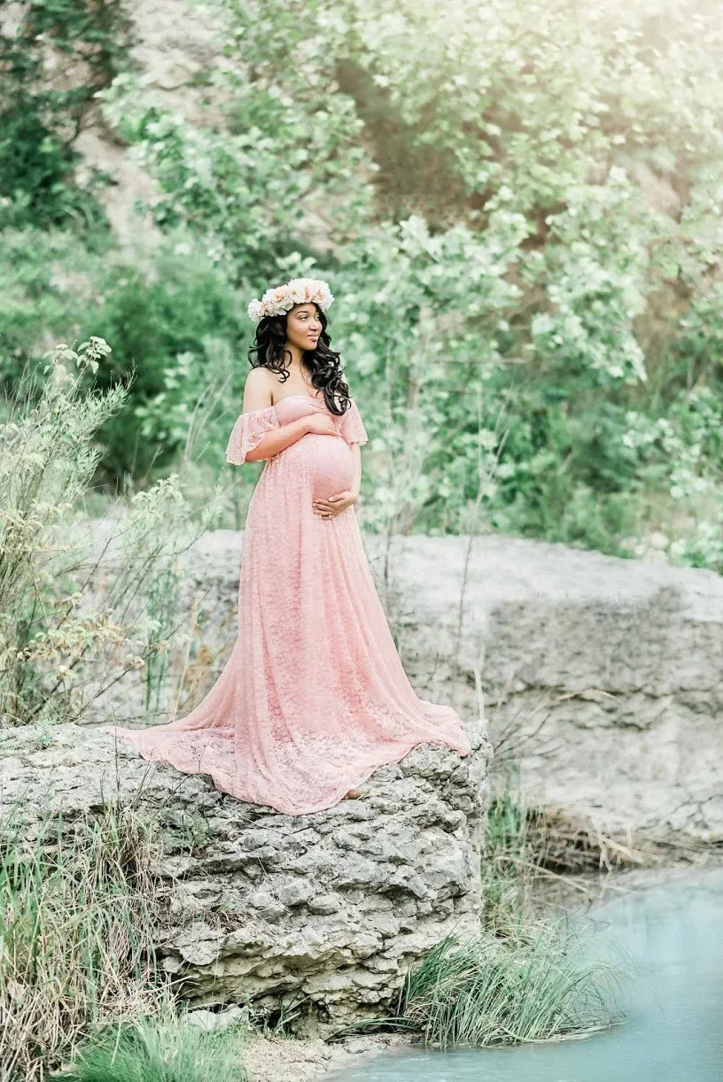 Pregnant woman in a pink dress standing in a natural setting with greenery and water.